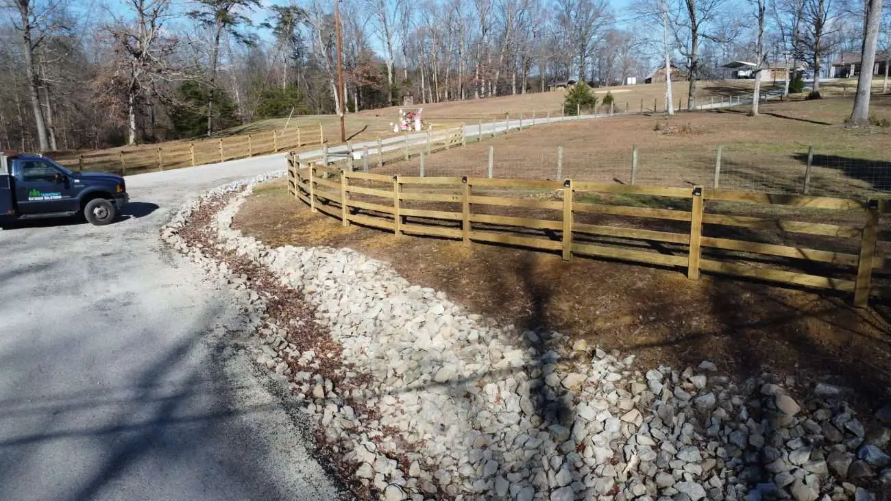 Wooden 4-board split-rail fence with natural drainage system and decorative rock along a rural driveway in Shepherdsville, Kentucky. Designed and installed by TerraCraft Outdoor Solutions to enhance property boundaries and improve stormwater management.
